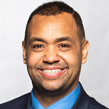 Man smiling brightly, wearing a blazer and blue collared shirt.