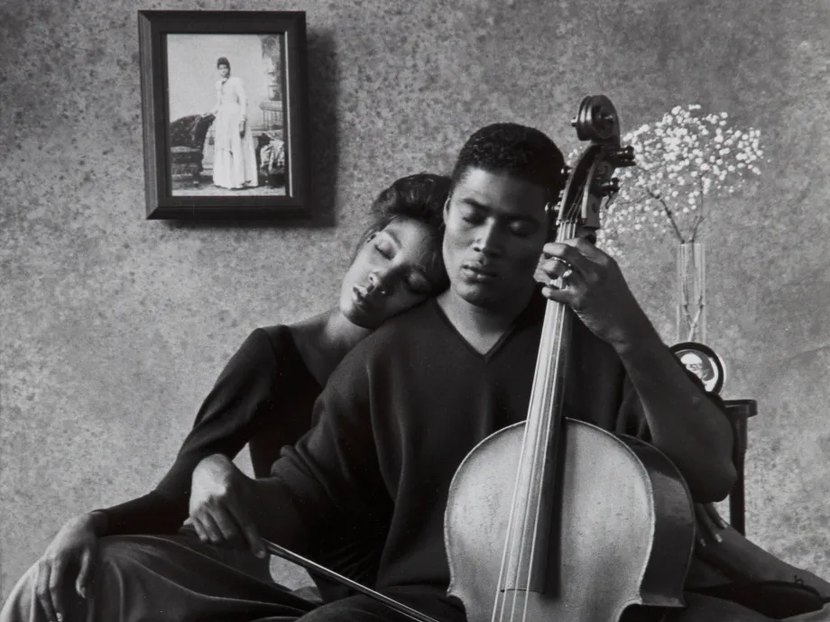 Black-and-white photograph of a man playing the cello while his wife leans on him