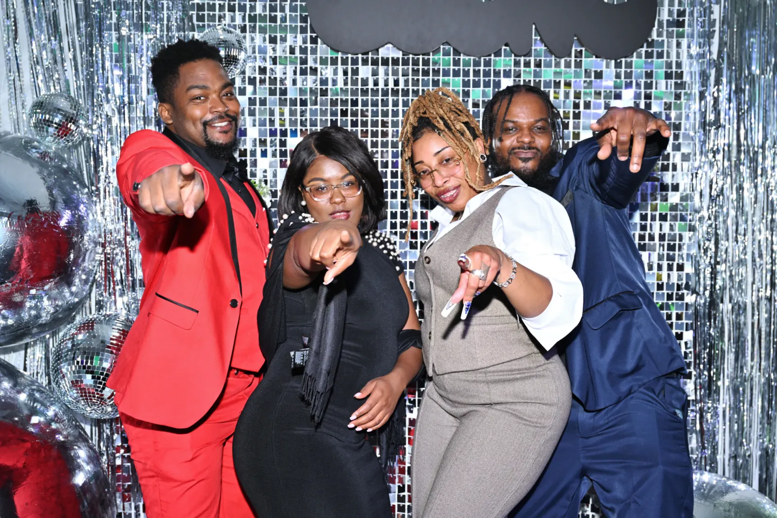 A group of 4 well-dressed people smiling and pointing at the camera against a discoball-like background.